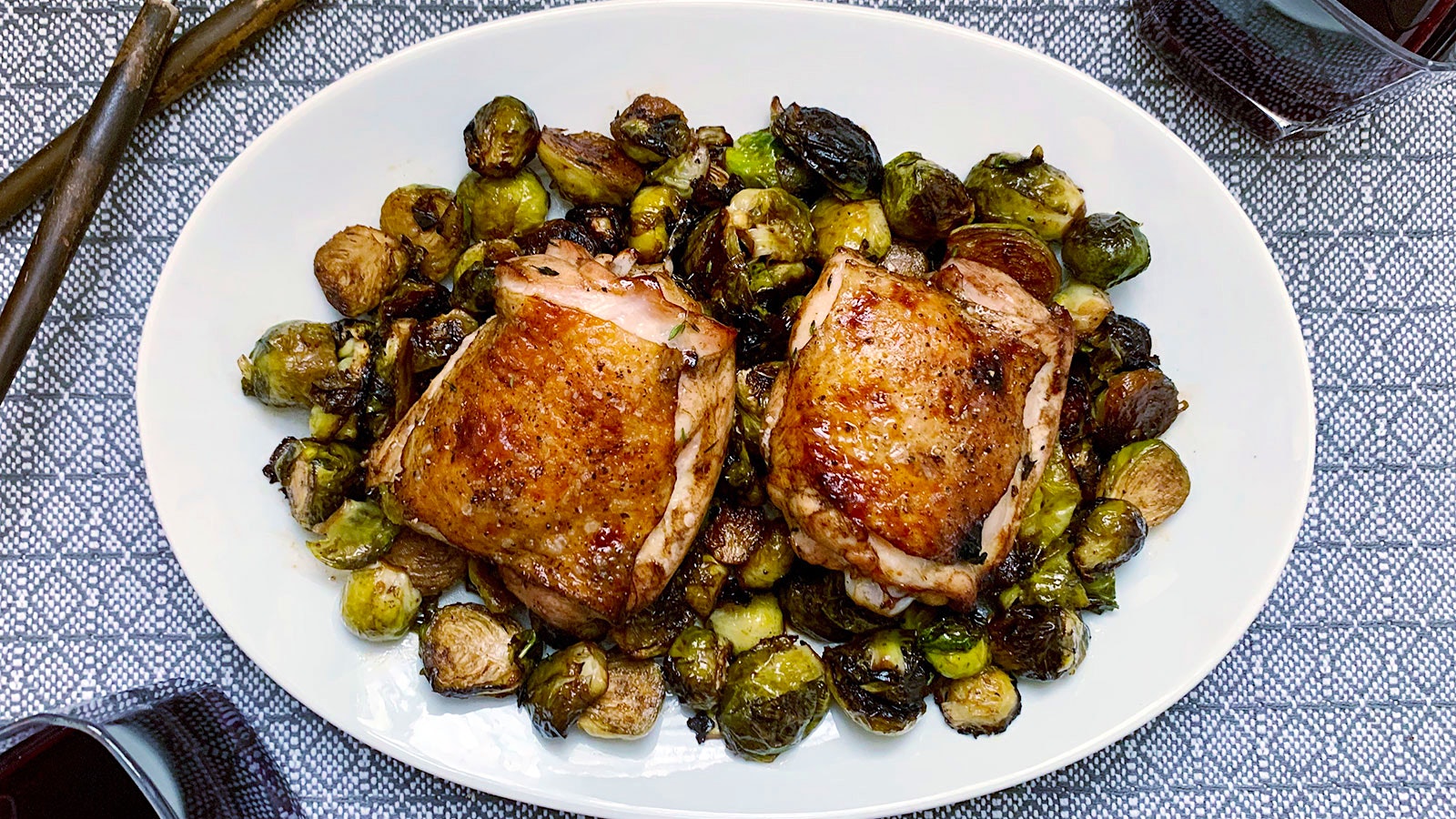  Two crispy-skinned chicken thighs on a white plate with balsamic-roasted brussels sprouts 